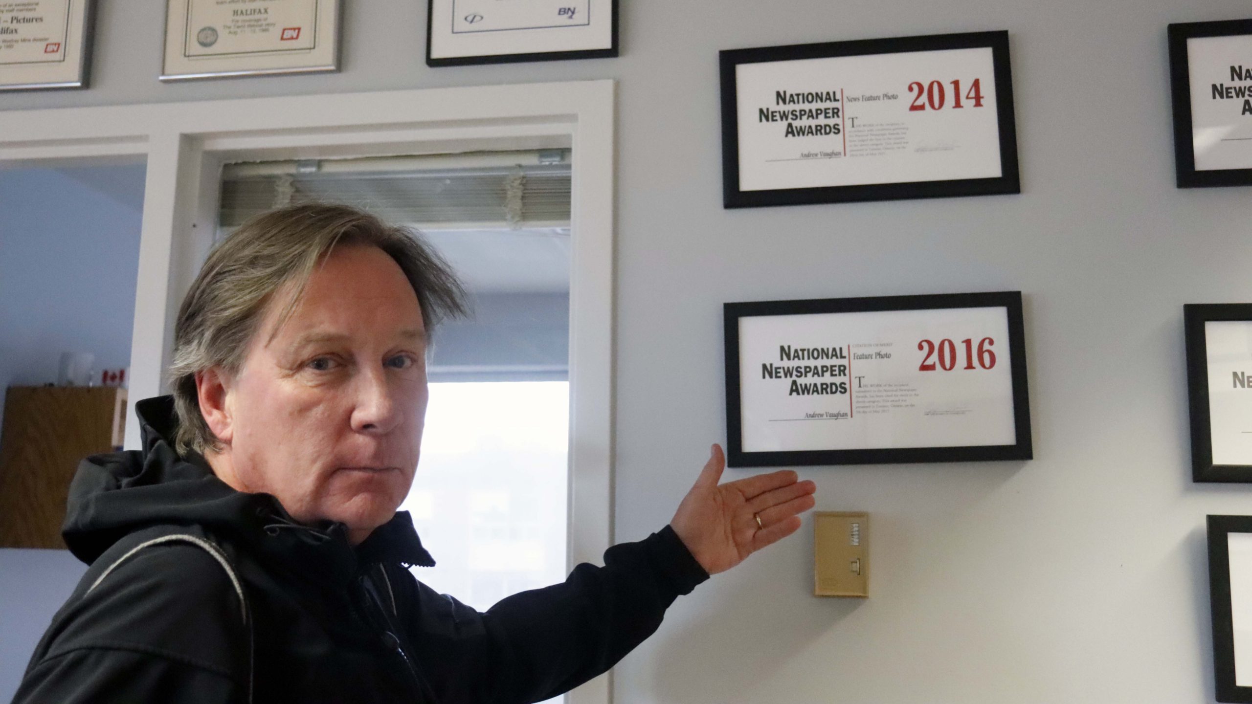 Andrew Vaughan has his hand up next to a plaque with the 2016 nomination for a National Newspaper Award, looking back to the camera.