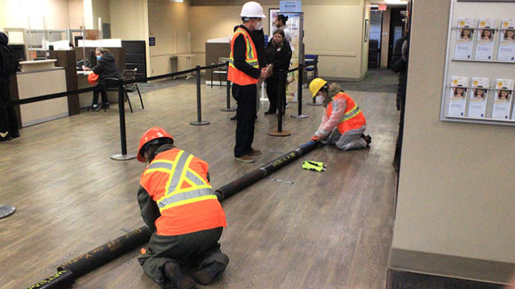 Two people in construction uniforms tape a pipeline to the ground in RBC. One person in a similar uniform stands and speaks to the onlookers. One person standing in line to speak with a teller watches the pipeline being installed.