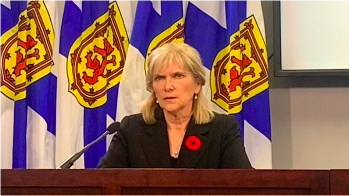 The Auditor General, Kim Adair, sits behind a mahogany desk wearing a black blazer and a poppy. Behind her are four Nova Scotia flags draped downwards with the crest in the center emphasized.