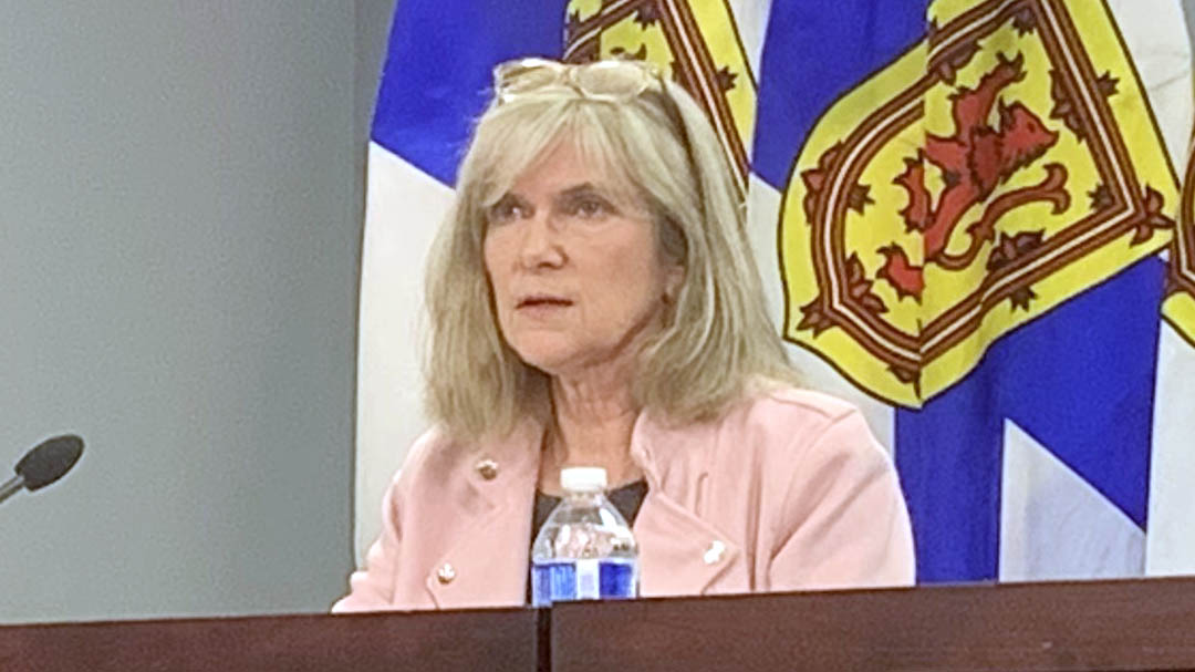 The Auditor General, Kim Adair, sits behind a mahogany desk wearing a light pink blazer. Behind her are four Nova Scotia flags draped downwards with the crest in the center emphasized.