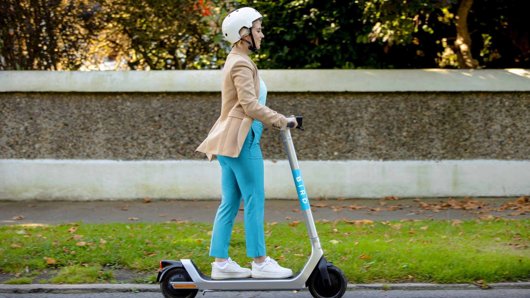 Side profile, full shot of a woman with a white helmet, bright blue pants and a brown coat riding a Bird Canada electric scooter over pavement on a sunny day. Green grass, a garden wall, and trees are in the background.