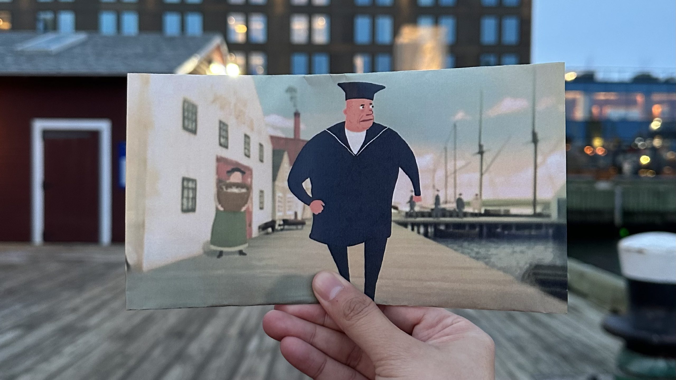 A printed still shot from The Flying Sailor, of a blue-suited sailor walking toward the viewer down the Halifax pier, held up by a hand in front of the real-life modern day pier, with the buildings and docks in the print lined up with the real-life surroundings so it appears to be one image.