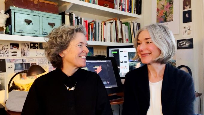 Amanda Forbis (left, with curly grey hair) and Wendy Tilby (right, with straight silver hair) look at each other, laughing, in front of a shelf and desk which holds books, a computer, lamp, small set of drawers, and still images from their animated work.