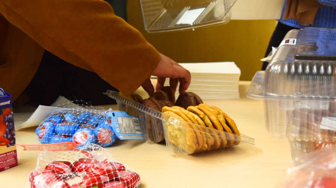 Hand holds reaches for chocolate chip cookie set on a table surrounded by snacks like cheese, croissants, and apples.