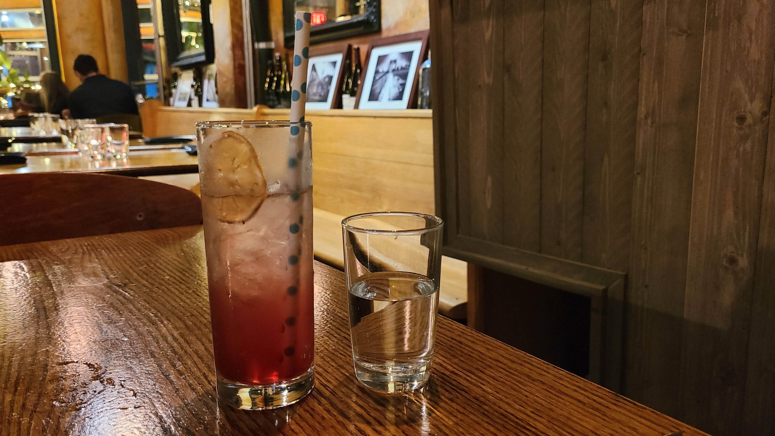 A glass containing a mocktail sits on a restaurant table to the left of a glass of water. The mocktail is garnished with a slice of dried fruit.