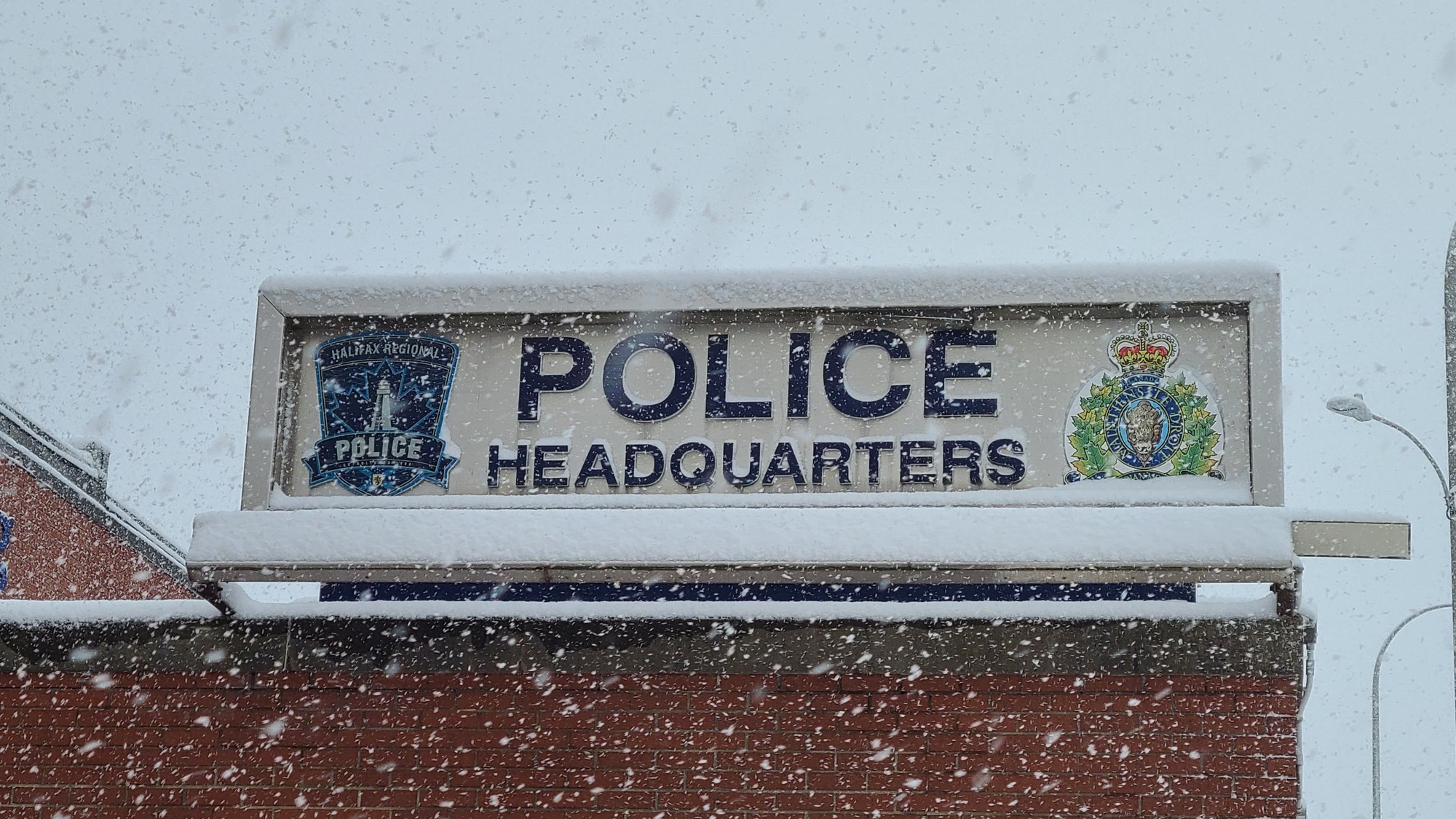 The sign outside of the Halifax Regional Police headquarters on Gottingen Street has the words police headquarters in the middle, the Halifax Regional Police crest on the left, and the Royal Canadian Mounted Police crest on the right. It is snowing and there is snow on the sign.