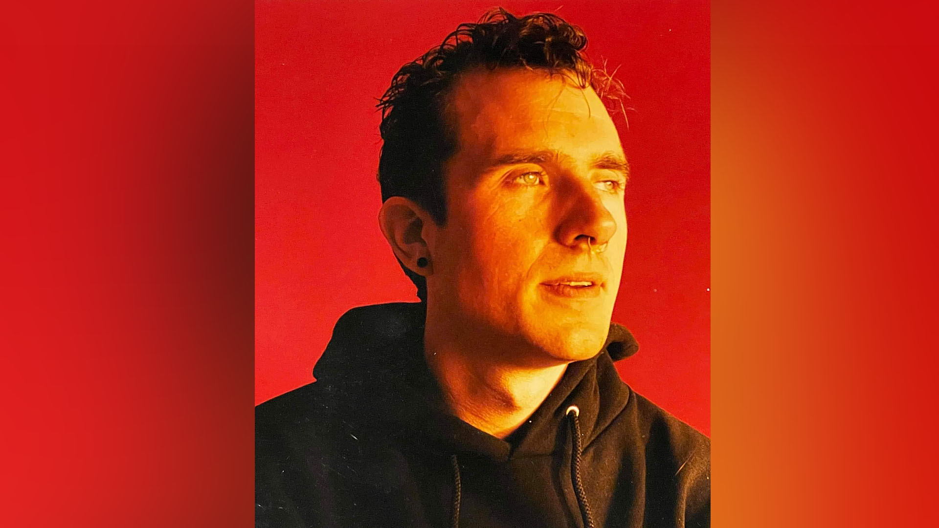 Chris Redden poses in front of a red backdrop. The photo is a close-up of his head and shoulders. He is wearing a black hoodie and one stud earring. He also has a nose ring. He is staring out into the distance.