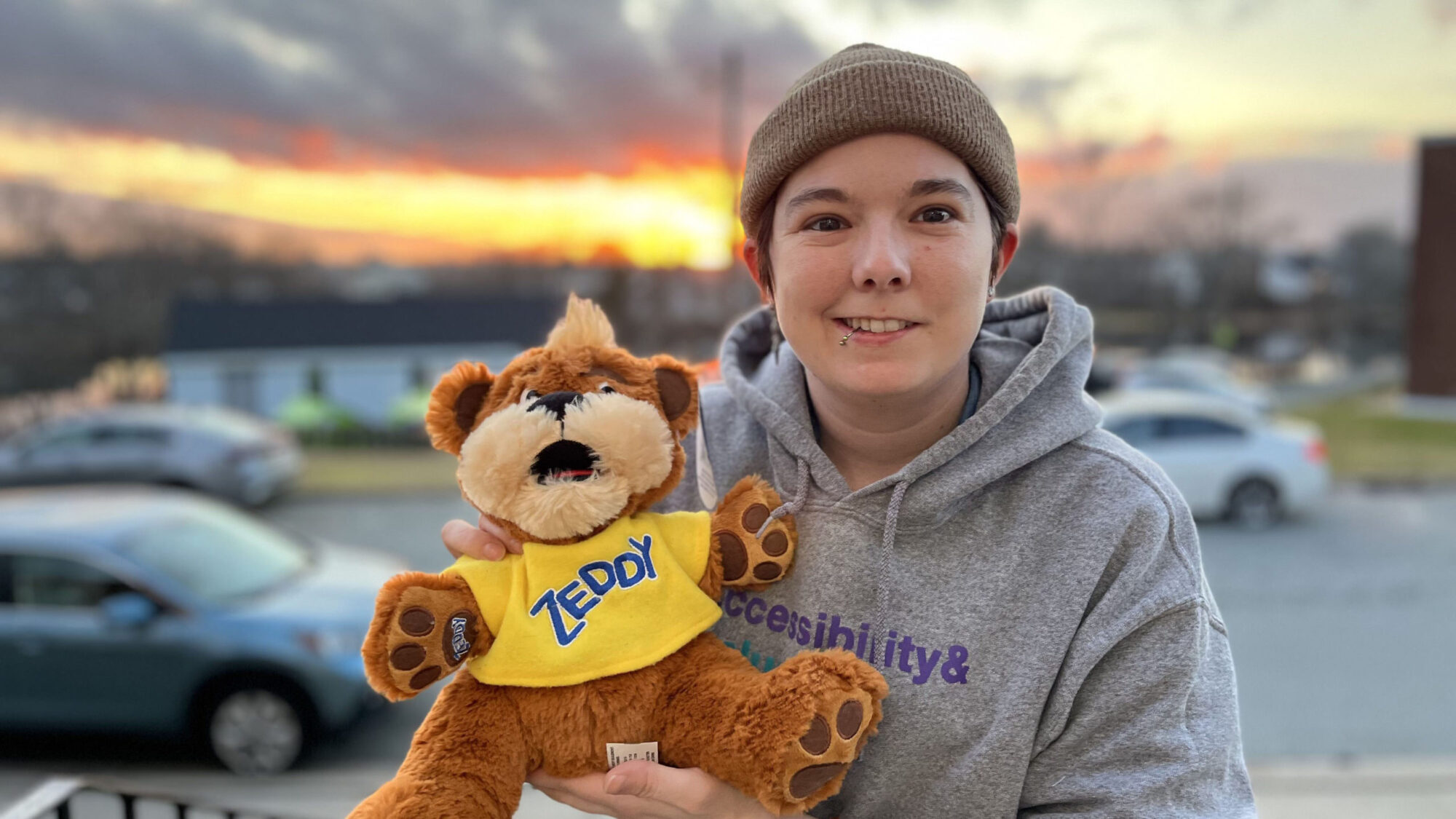 Nat Cook is holding up their Zeddy with a sunset behind them. Cook is wearing a grey hoodie and brown beanie. The bear is orange-brown with a yellow T-shirt on that says: 'ZEDDY' in blue letters.