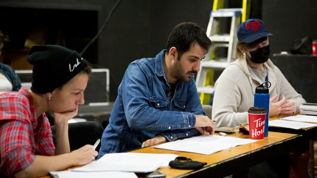 Three people sit at a table filled with pages listening to actors read at Halifax’s OutFest workshop in April 2022. From left to right, Dramaturg Annie Valentina wears a black toque and red flannel. Artistic director Isaac Mulè sits in the middle turning a page while wearing a denim, button-down shirt. On the right, McKenna James Boeckner wears a cream-coloured sweater and a blue baseball cap with the message “TUE U” written in red.