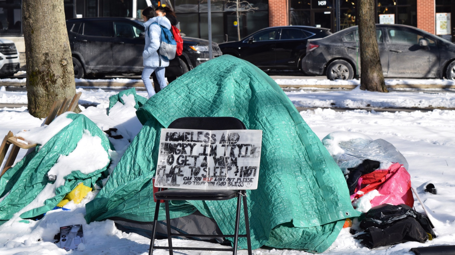 Tent covered in green tarp sits in the snow-covered park as pedestrians walk by on the pavement behind it. A white signed propped up on a black chair reads: “Homeless and hungry I’m trying to get a warm place to sleep & hot meal. Can you help a guy out please and thank you God bless.”