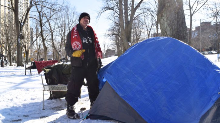 Man wearing black toque, black jacket and red scarf stands beside a grey tent with a blue tarp drapped on top of it in a snow-covered park. He wears one yellow glove and winter boots. There’s a metal chair behind him with clothes on it. 