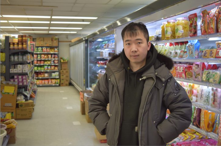 Man wearing a black hoodie and green winter jacket over top poses for a picture with his hands in his pocket. He stands inside a grocery store in front of a refrigerated section filled with colourful packages.