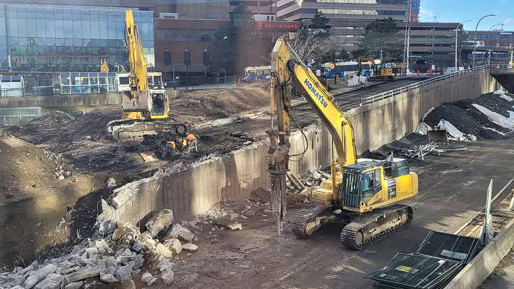 Two large excavators and three men work to deconstruct a large concrete wall in the middle of a city.
