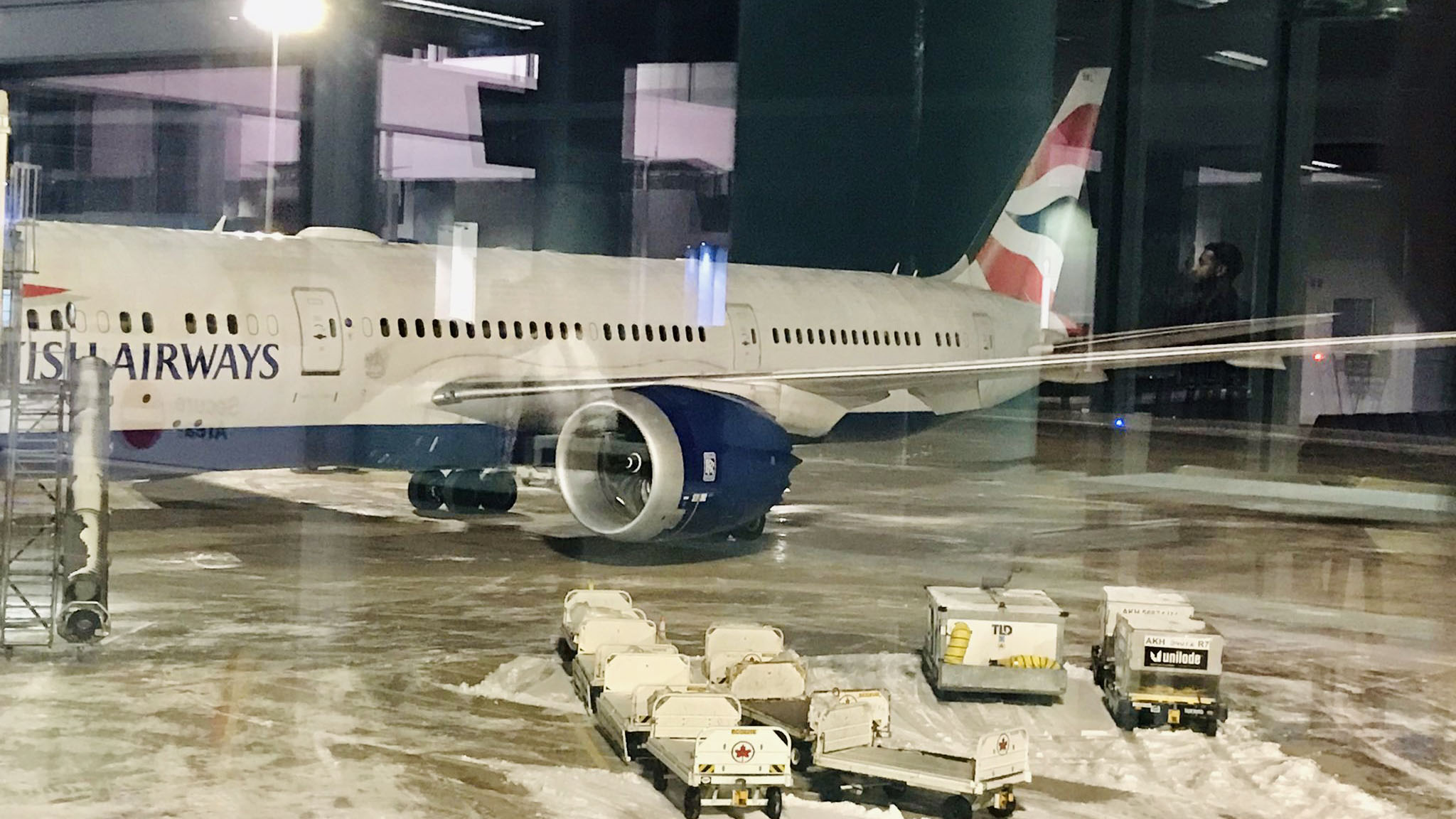 British Airways flight 216 sits at the arrival gate. It is dark outside and there is snow on the ground. The photo is taken through the window of the airport.