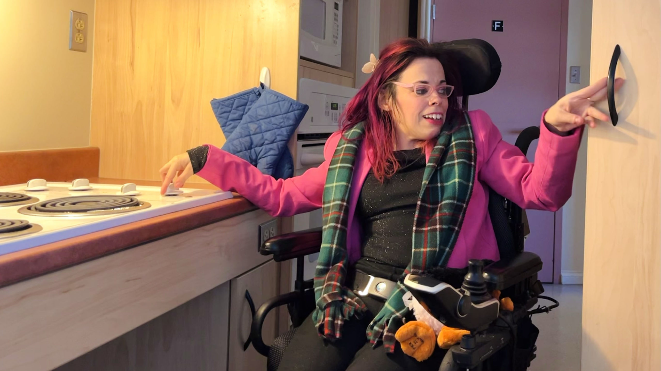 Carrie-Ann Bugden opens the cupboard and turns on the stove in her kitchen. She is in her wheelchair, wearing a pink blazer, black shirt, and Newfoundland scarf. In her lap is a puffin stuffed animal, representing her home province.