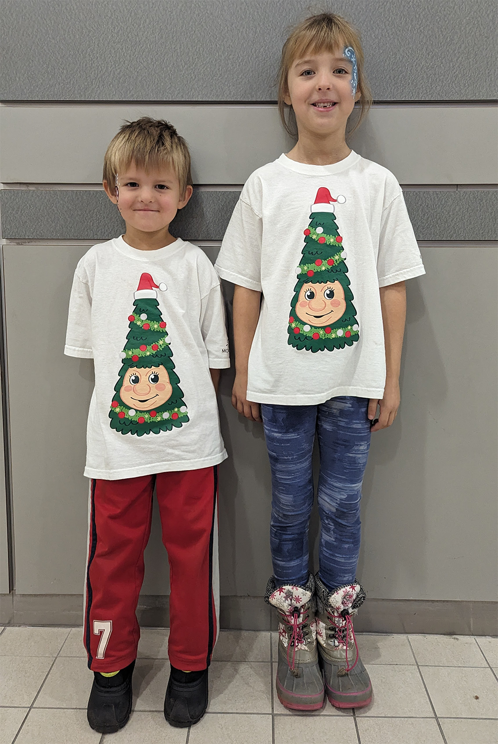 A young brother and sister stand in front of a grey wall smiling at the camera with Christmas inspired face paint. They wear white T-shirts with Woody the Talking Christmas Tree on the front.