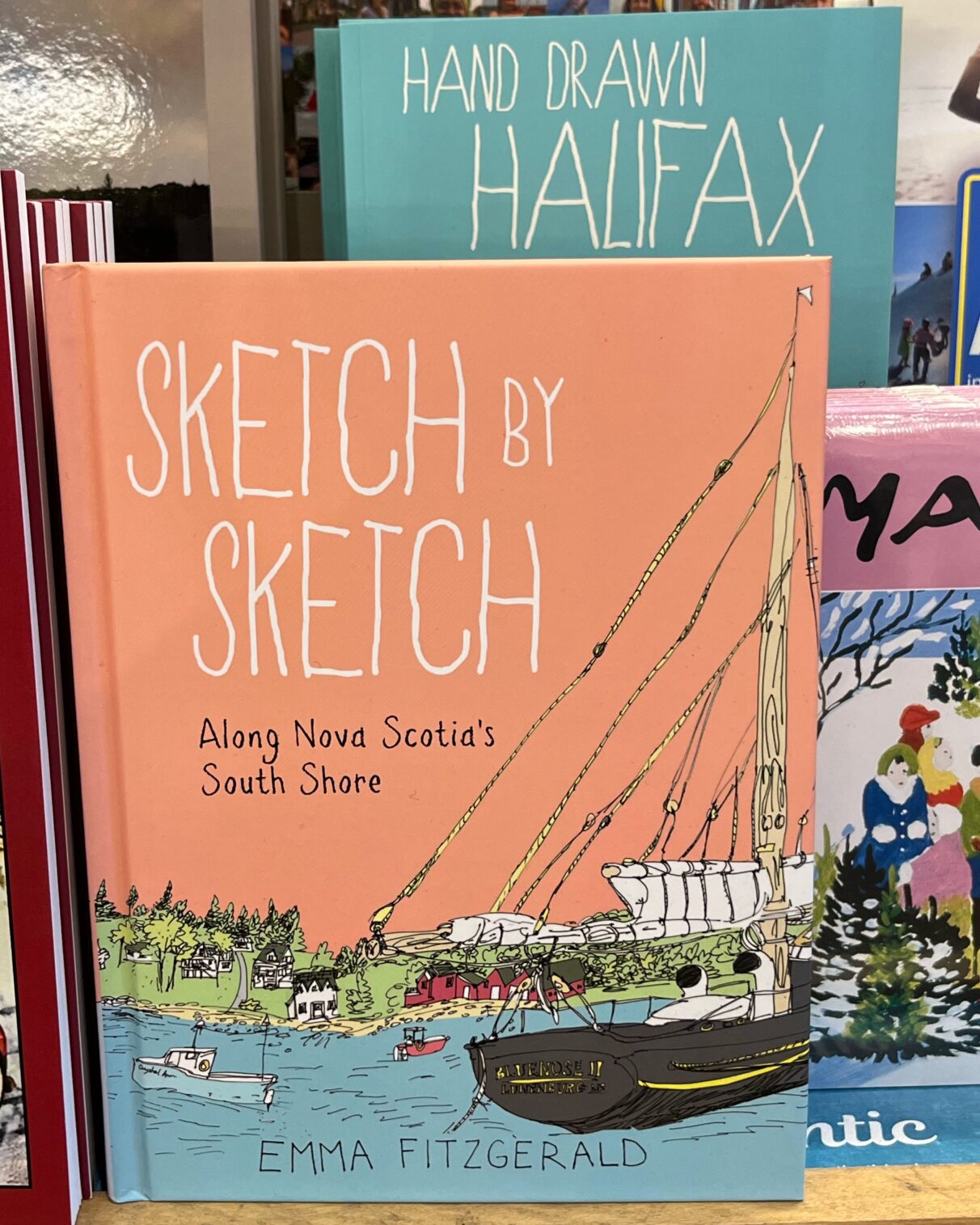 Emma FitzGerald's book, 'Sketch by Sketch' is on the counter at a book store. The book has a picture of the Bluenose on the cover. Behind is her book Hand Drawn Halifax.