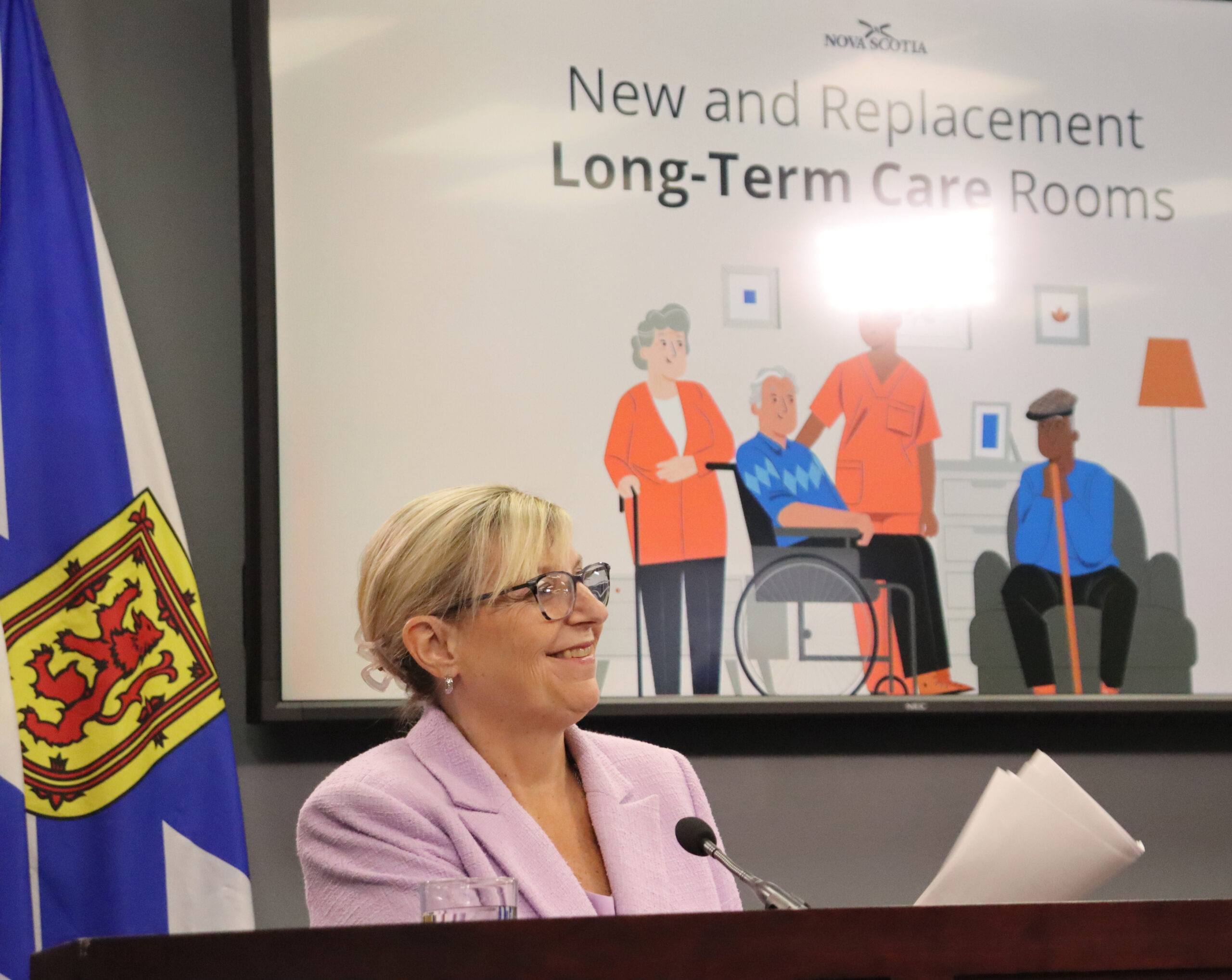 A woman smiling in front of a projector that has the words "New and Replacement Long-Term Care Rooms"