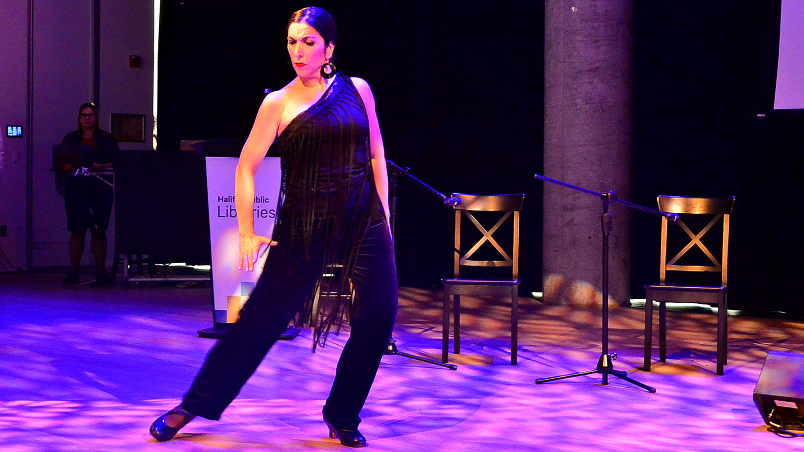 A lady dressed in a black jumpsuit with fringes dances to flamenco music.