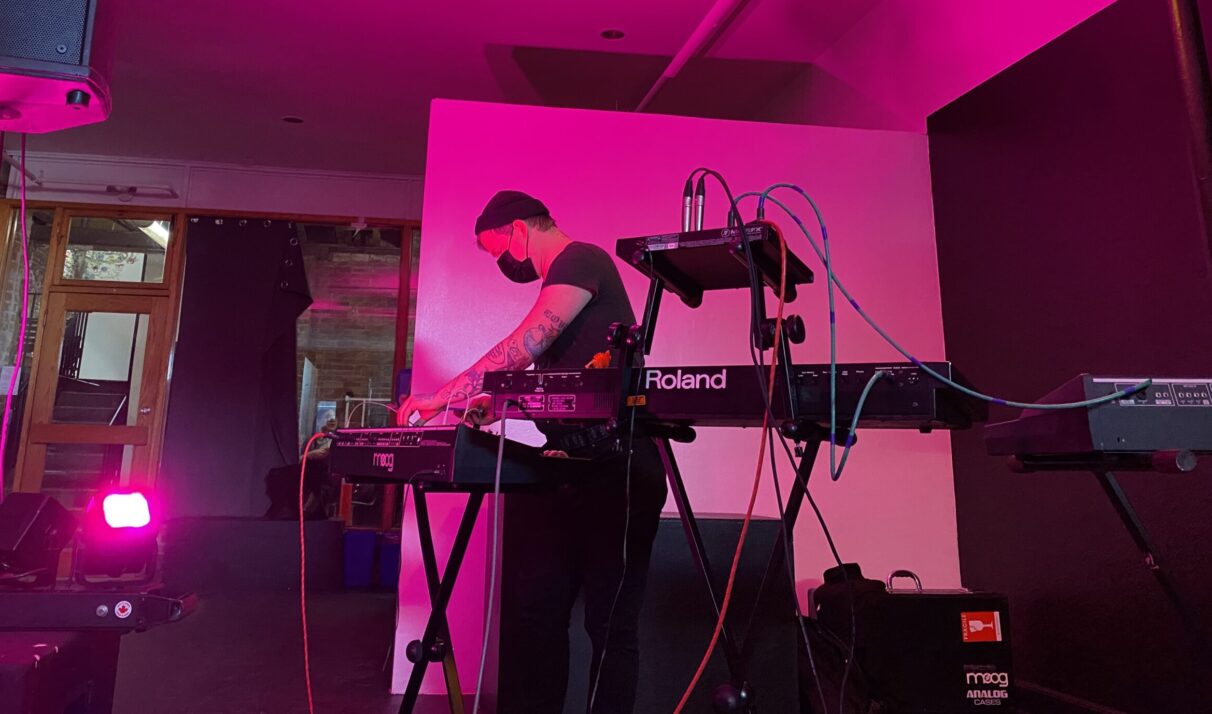 Woman wearing a beanie performing with three synthesizers under pink light