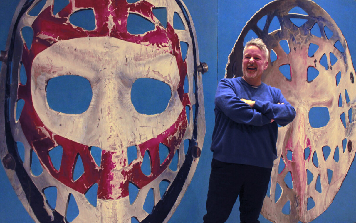 A laughing man standing front of a painting of hockey mask.