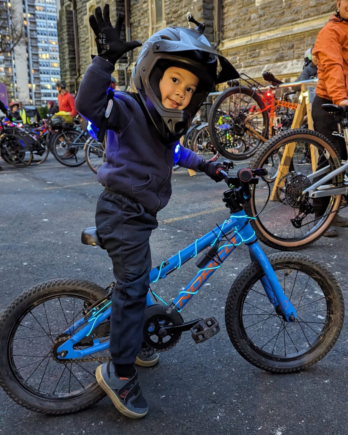 Side photo of a little boy smiling at the camera. He's sitting on his blue bike with his feet on the ground. His right hand is in the air.