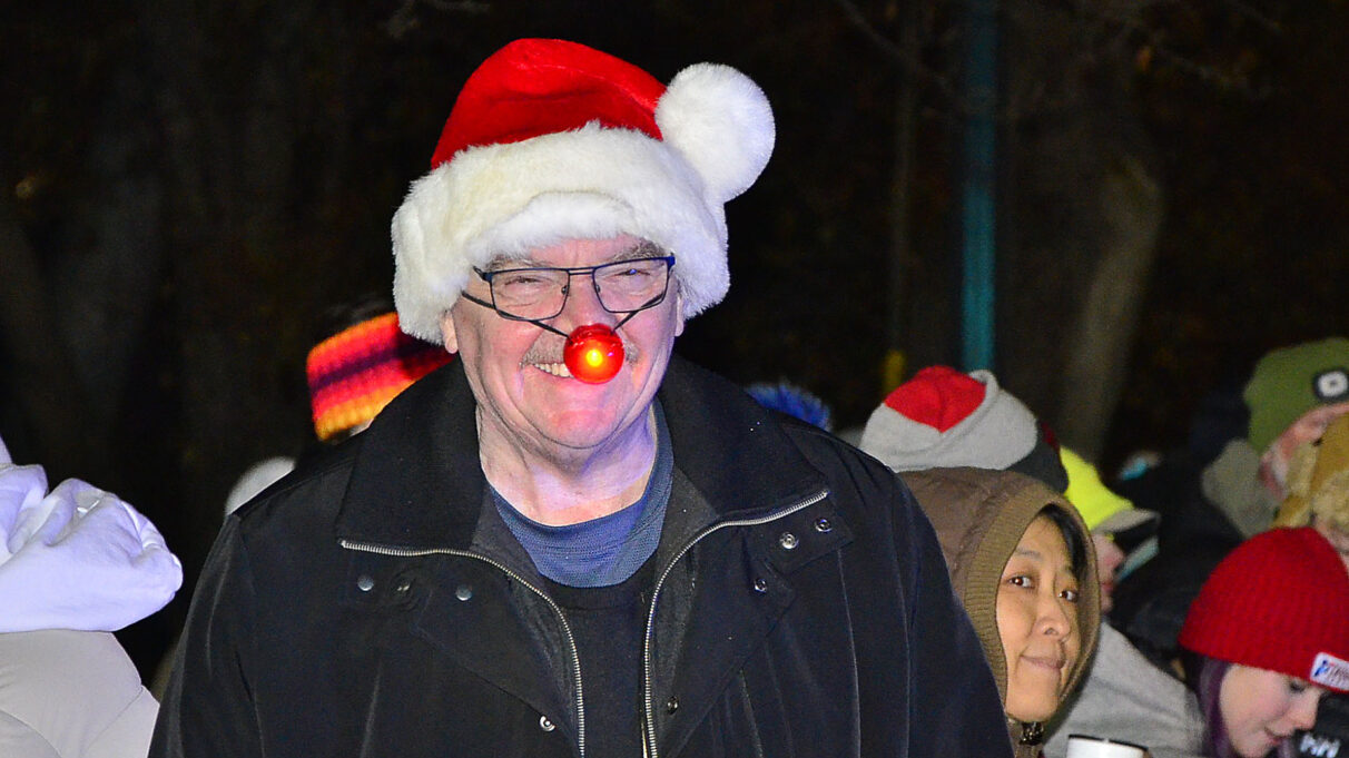 A man is smiling while wearing a Santa hat and a red glowing Rudolph nose.
