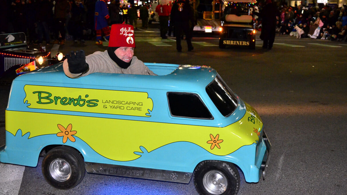 A little car, painted like the Mystery Machine, is driven by a man wearing a red fez hat waving to the crowd.