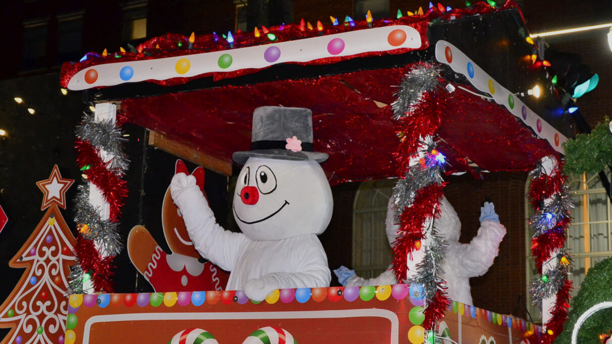 Frosty the Snowman waves at the crowd while he is on a colourful float, lined with tinsel and lights.