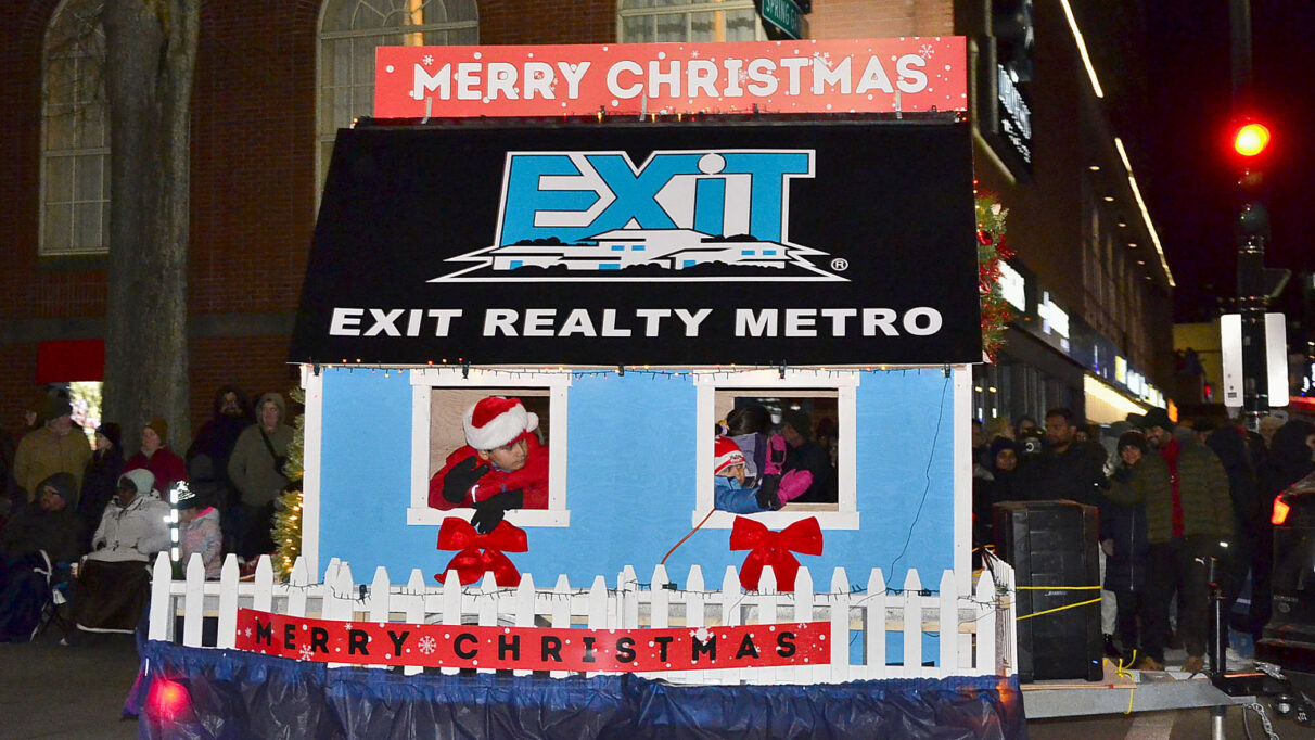 An Exit Realty parade float of a miniature blue house with a white picket fence has a Merry Christmas sign hung on the front.