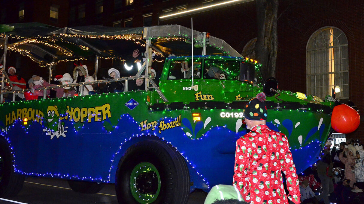 The green and blue Harbour Hopper was covered in lights and a red Rudolph nose. A man dressed in a red suit with snowmen covering it walks beside the vehicle.