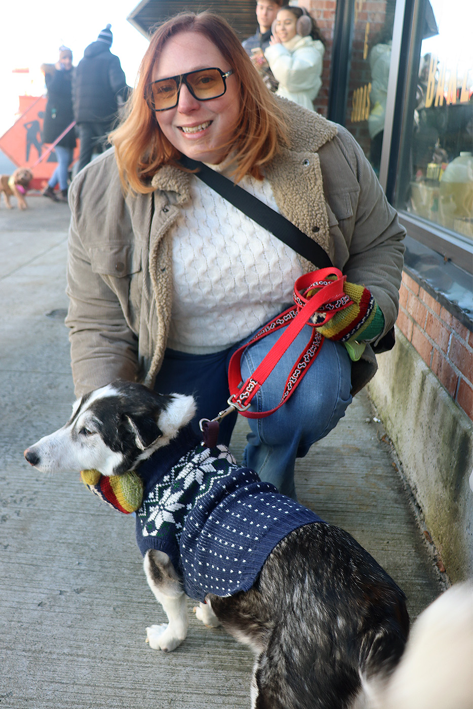 A woman kneels down beside her black and white dog wearing a blue sweater.