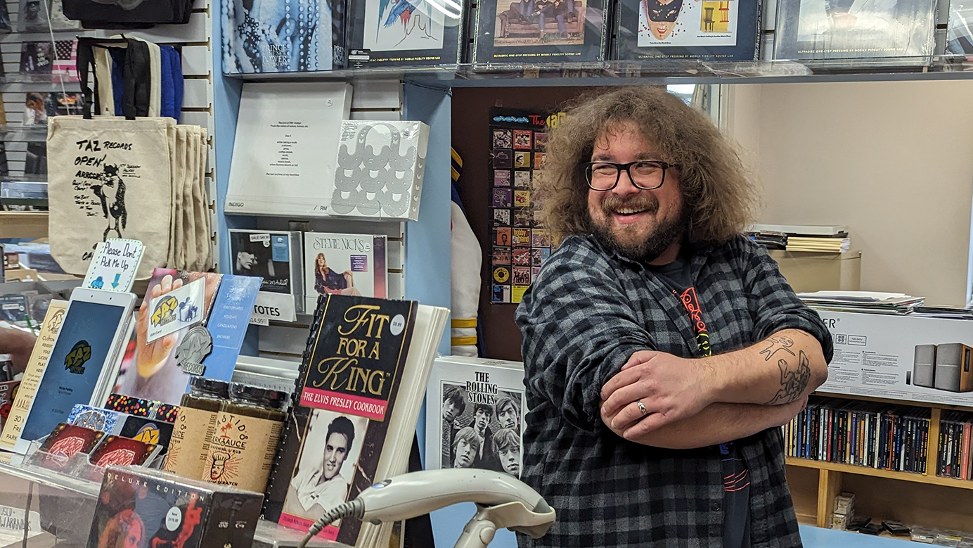 A man with frizzy shoulder length shares a laugh behind the cash desk as he crosses his arms at Taz Records store in Halifax.