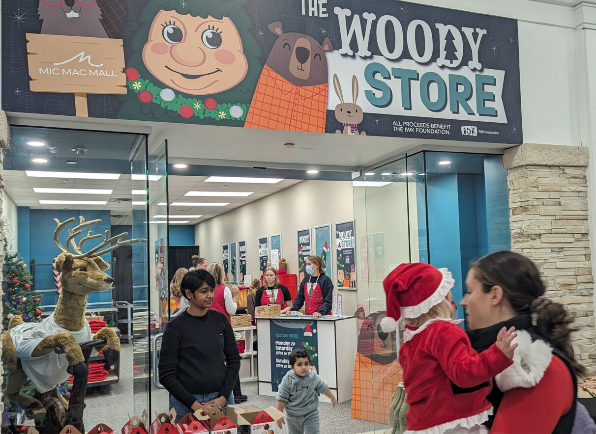 A woman carrying a young girl dressed in a Santa outfit stands in front of the new Woody merchandise store. A woman stands in front of the glass doors with a table full of Timbit boxes to hand out individual Timbits to customers as they leave and/or enter the store.