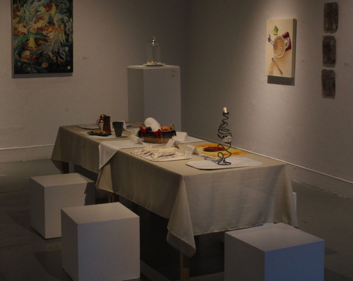 A table with crafts at its six table settings sits in the middle of a gallery, surrounded by two paintings on the wall and a podium.
