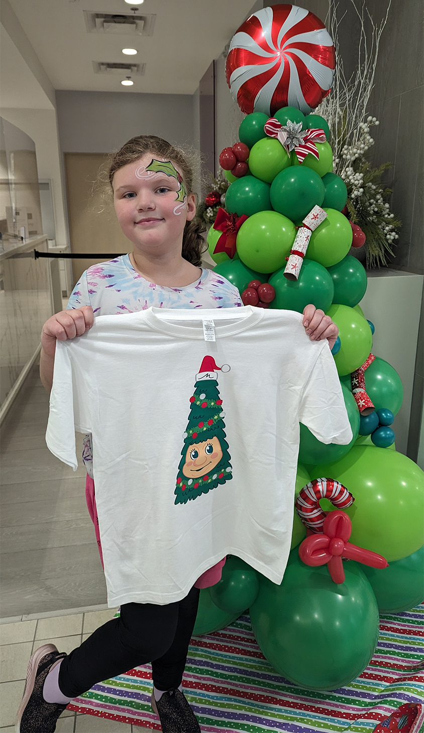 A young girl with a painted Christmas holly on her face holds a white T-shirt with Woody the Talking Christmas Tree on the front of it. She stands in front of a balloon Christmas tree.