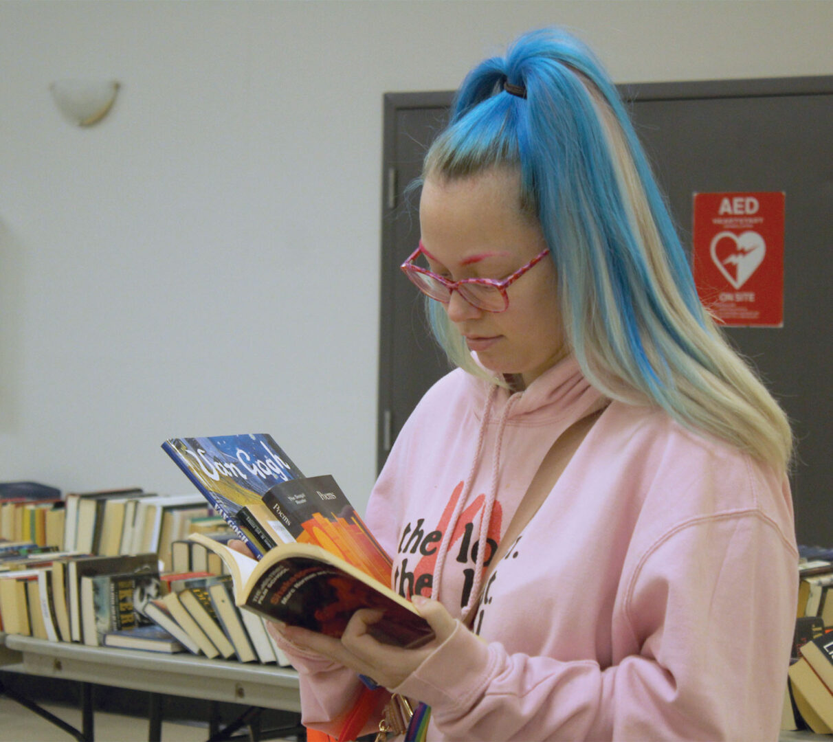 A young blonde woman with streaks of dyed-blue hair standing in front of a table of books, while flips through a book in her hand, while holding other books between her arm.