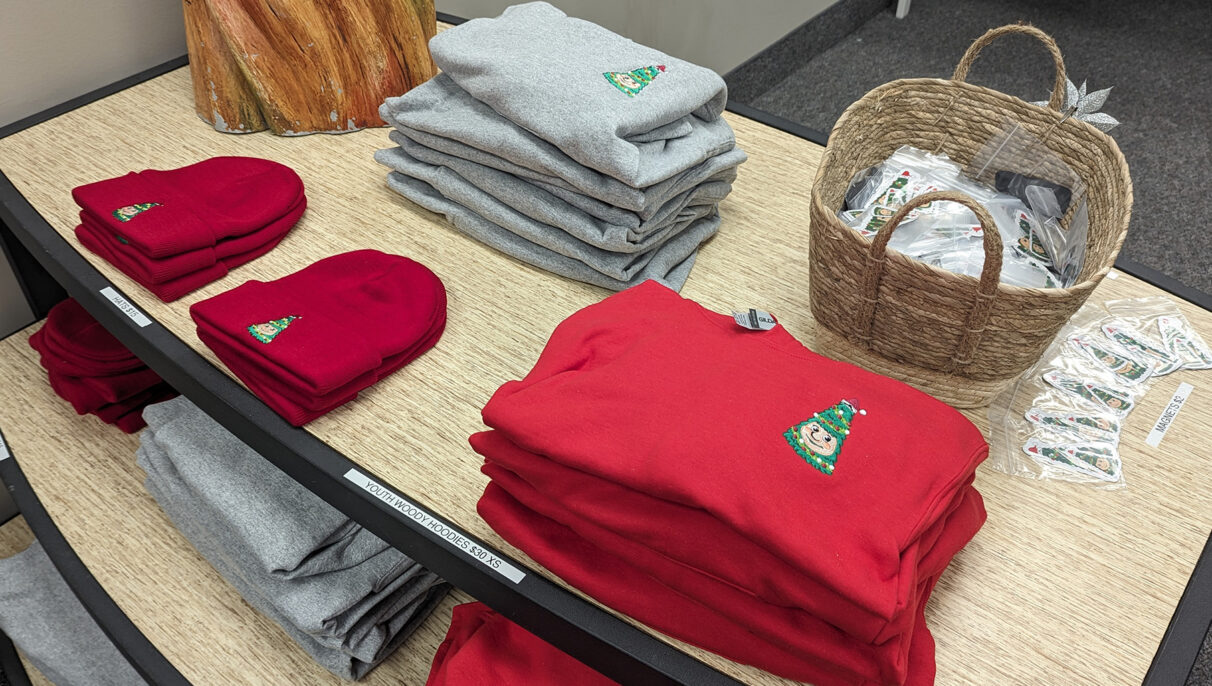 Red crew necks, red hats and grey sweaters with Woody the Talking Christmas Tree logo on the front. a basket full of Woody the Talking Christmas Tree magnets.