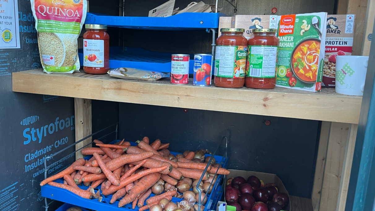 Interior of the food pantry at the Dartmouth Community Fridge. The top shelf displays canned items and the lower shelf is filled with carrots, onions and apples.
