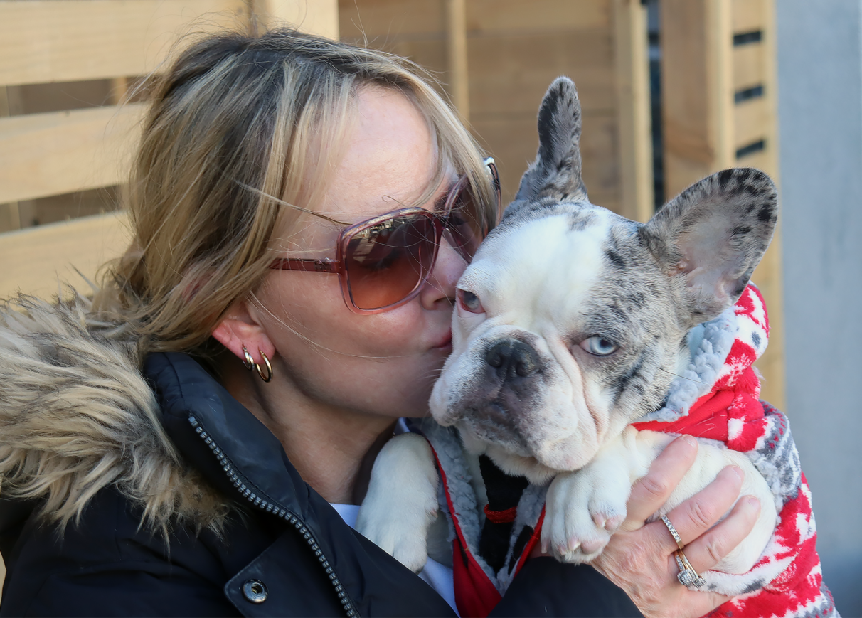 A woman wearing sunglasses gives her french bulldog a kiss on the cheek.