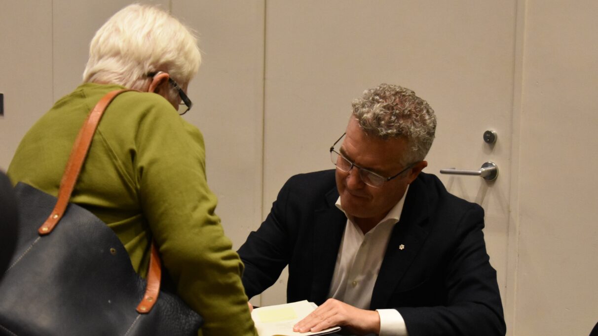 A woman in a green shirt with her back to the camera on the left, and a man facing the camera in a white dress shirt and black jacket is signing a book on the right.
