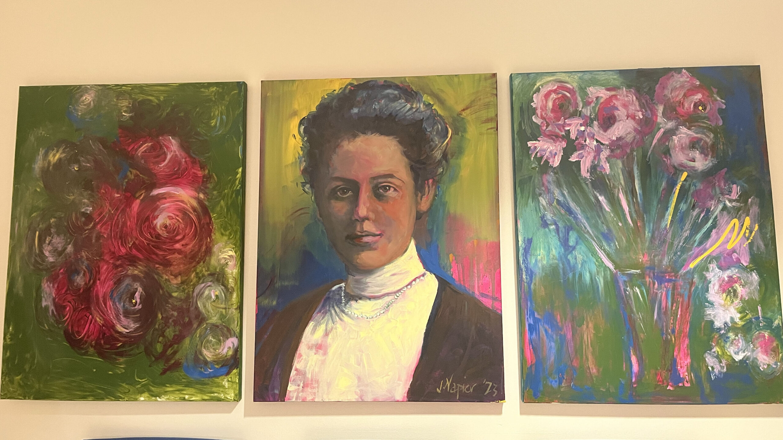 Three portraits are hanging on a wall. The portrait in the middle is of a woman with dark hair, wearing pearls. The other two are of flowers that are red and pink.