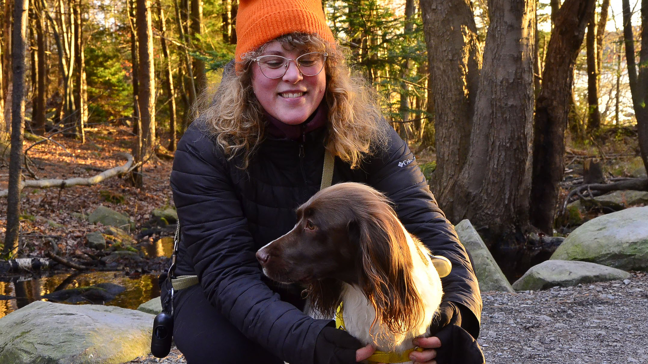A woman with curly blonde hair, glasses and an orange hat has her arms around a brown and white dog.