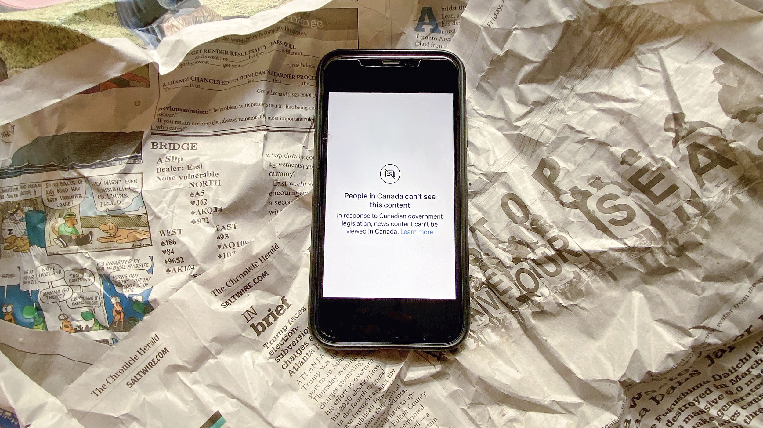 A phone with a blocked news site sits on a crumpled up newspaper.