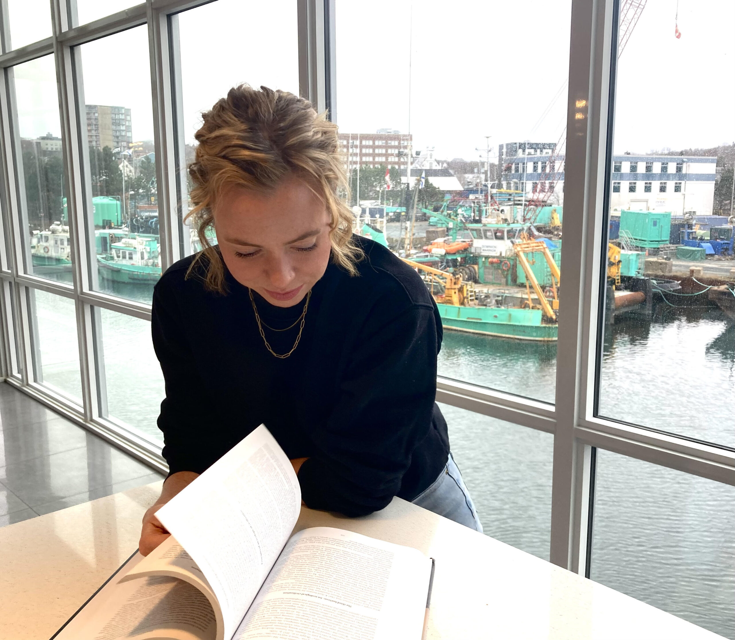 Woman with blonde hair reads textbook in room on the ocean with boats in the background