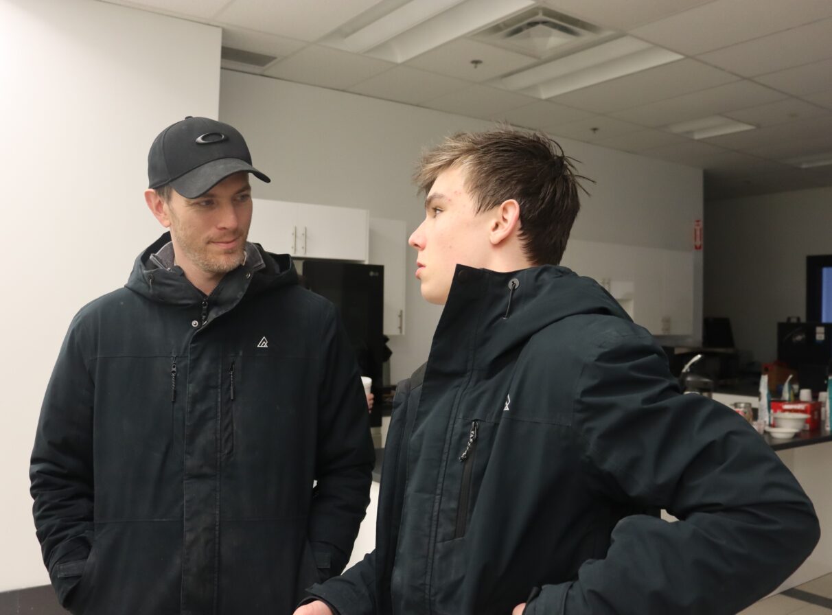 A man in black clothes and a black hat looks at a talking boy in black clothes.