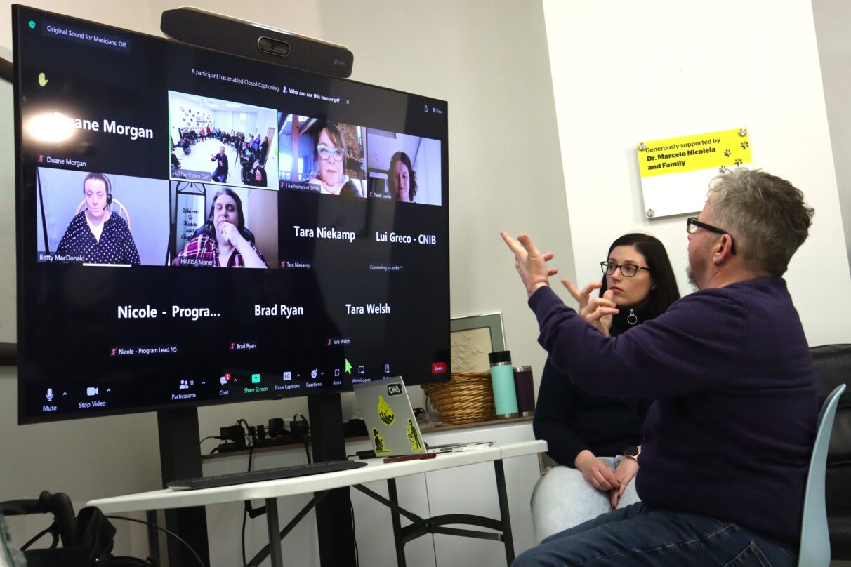 A man gestures in front of the screen. A woman sits beside the screen. On the screen is the Zoom meeting interface.