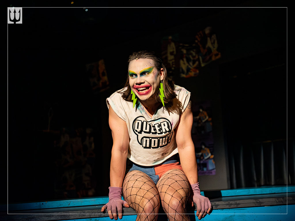 Actor sits with a painted face in the spotlight on stage.