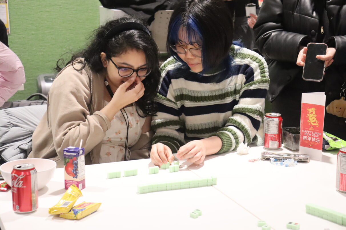 A girl in a striped sweater is teaching another girl to play mahjong.