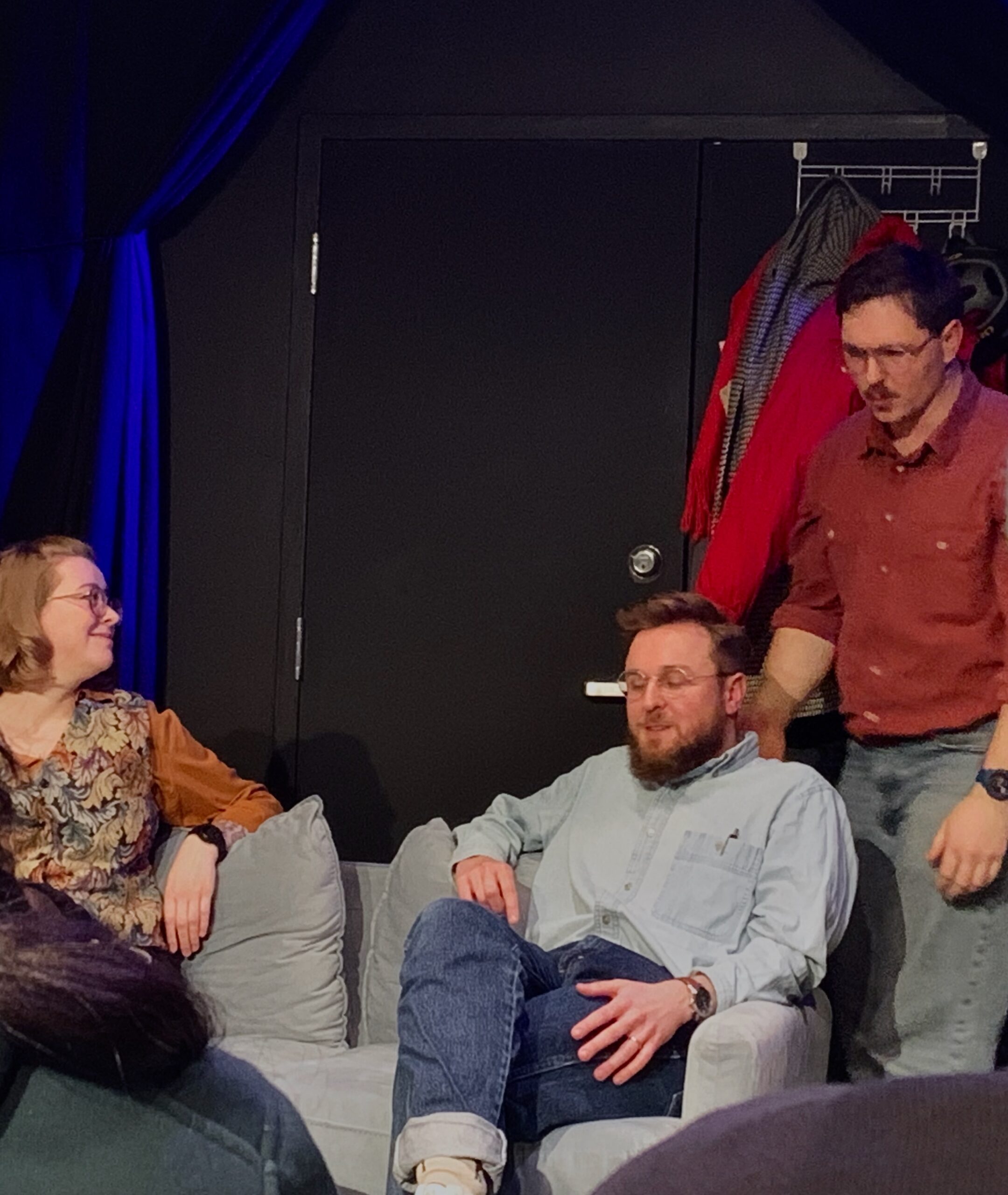 On a stage, two actors sit on a couch while one stands.
